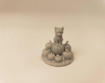 Puede incluir: Una pequeña figura de color gris monocromático que muestra a un gato sentado sobre una calabaza central, rodeado de varias calabazas más pequeñas dispuestas sobre una base circular. La escultura tiene un color sólido con texturas detalladas.