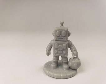 Puede incluir: Una pequeña figura de robot gris monocromática de pie sobre una base circular con baldosas. El robot presenta una cabeza cuadrada con dos ojos redondos, una pequeña antena en la parte superior y un torso cuadrado con varios botones. Sostiene un cubo redondo.