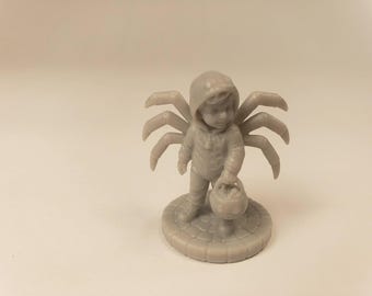 Puede incluir: Una figura impresa en 3D de color gris claro que representa a un niño con un disfraz de araña con capucha y ocho patas que se extienden desde la espalda. La figura se apoya sobre una base circular de adoquines y sostiene un cubo.