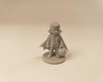 Puede incluir: Una pequeña figura gris monocromática de un niño disfrazado de vampiro, con una capa de cuello alto, traje y pajarita. La figura se apoya sobre una base circular y sostiene en una mano un pequeño cubo con forma de calabaza.