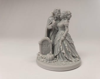 Puede incluir: Una figura de resina gris que representa a un vampiro con capa y a una persona con un vestido con volantes de pie. Junto a ellos hay una lápida con el texto RIP y un corazón, además de una pequeña calabaza y hojas sobre una base circular.