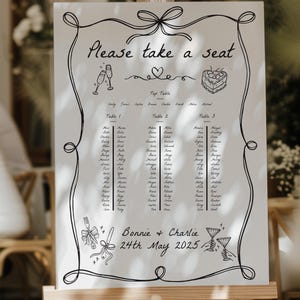 May include: A white wedding seating plan displayed on an easel, with black handwritten text and decorative line art. The text includes "Please take a seat," names, and the date "24th May 2025." The design incorporates bow and ribbon motifs.