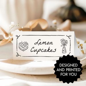 May include: A white place card featuring the words "Lemon Cupcakes" in black script, accompanied by a cake and flower illustration. The card is decorated with black flourishes and the text "DESIGNED AND PRINTED FOR YOU" in a black starburst.