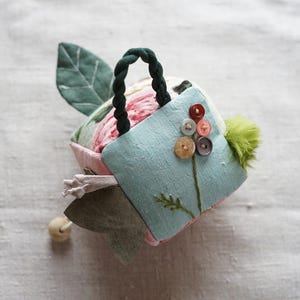 Può includere: Un piccolo cuscino per spilli in tessuto fatto a mano a forma di casa con un tetto di foglie verdi, una porta blu e un fiore fatto di bottoni sul davanti.