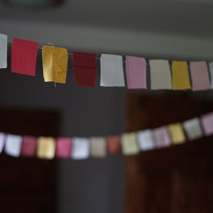 May include: A garland of small fabric squares in various colours hangs from a wire. The squares are in shades of white, red, yellow, and pink. The background is blurred, with a dark interior and wooden doors.
