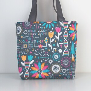 Może przedstawiać: Szara torba typu tote z kolorowym wzorem kwiatów, symboli matematycznych i kodu binarnego. Tekst na torbie brzmi: "F=ma", "V=IR", "π" i "01010011 01010100".