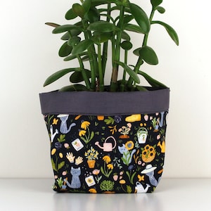 May include: A green jade plant in a black fabric planter with a colorful pattern of cats, flowers, and gardening tools.