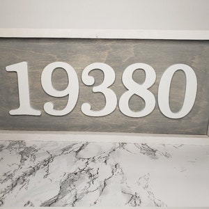 May include: A rectangular wooden sign displaying the number "19380" in white against a grey backdrop. The sign is framed in white. A stylish piece for home decor or as a house number, with a modern design.