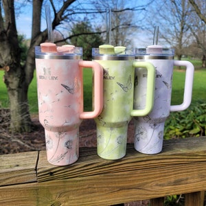 May include: Three Stanley tumblers with handles and straws. The tumblers are pastel pink, green, and lavender, each with a matching handle and lid. They feature a butterfly and dandelion design. The Stanley logo is visible on each tumbler.