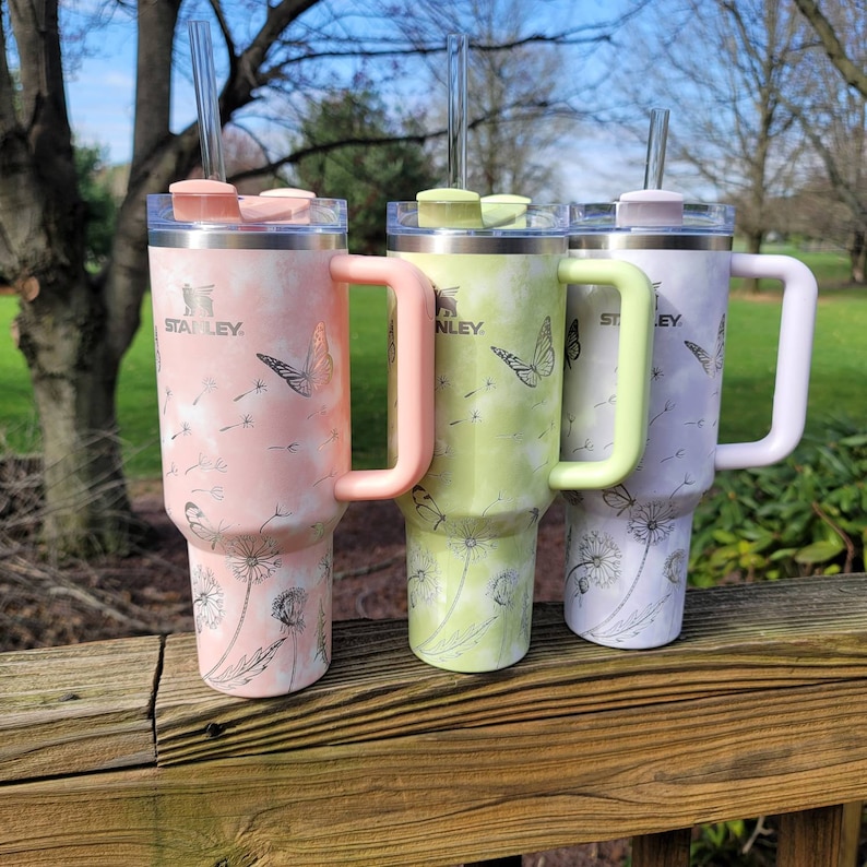 May include: Three Stanley tumblers with handles and straws. The tumblers are pastel pink, green, and lavender, each with a matching handle and lid. They feature a butterfly and dandelion design. The Stanley logo is visible on each tumbler.