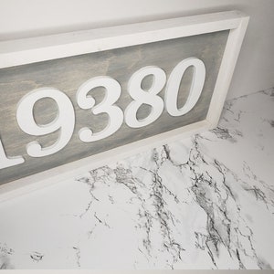 May include: A wooden address sign displaying the number "19380" in white, raised lettering. The background is grey-stained wood, framed by a white border. The sign is angled, showing the address against a marble-effect surface.