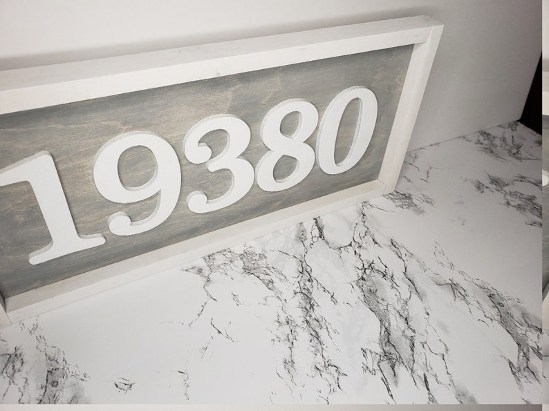 May include: A wooden address sign displaying the number "19380" in white, raised lettering. The background is grey-stained wood, framed by a white border. The sign is angled, showing the address against a marble-effect surface.