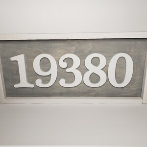 May include: A rectangular wooden sign displaying the number "19380" in white against a grey backdrop. The sign is framed in white, offering a clean, contemporary look. The numbers are raised, providing a three-dimensional effect.