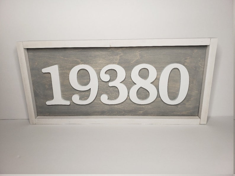 May include: A rectangular wooden sign displaying the number "19380" in white against a grey backdrop. The sign is framed in white, offering a clean, contemporary look. The numbers are raised, providing a three-dimensional effect.