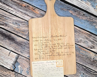 May include: A wooden cutting board with a handle, featuring handwritten recipe notes. The board is light brown with a smooth surface. The recipe card is attached to the board. The text on the board is in black ink. The board is on a wooden surface.