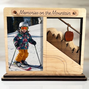 May include: Wooden photo frame with the text "Memories on the Mountain". The frame features a photo of a child skiing and a laser-cut mountain scene with a ski lift. The frame is made of light and dark wood.