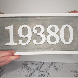 May include: A rectangular wooden sign displaying the number "19380" in white against a grey backdrop. The sign is framed in white. The sign is held by two hands.