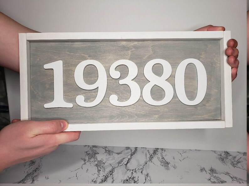 May include: A rectangular wooden sign displaying the number "19380" in white against a grey backdrop. The sign is framed in white. The sign is held by two hands.