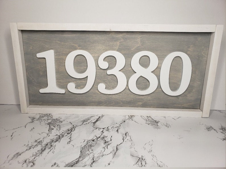May include: A rectangular wooden sign displaying the number "19380" in white against a grey backdrop. The sign is framed in white. A stylish piece for home decor or as a house number, with a modern design.