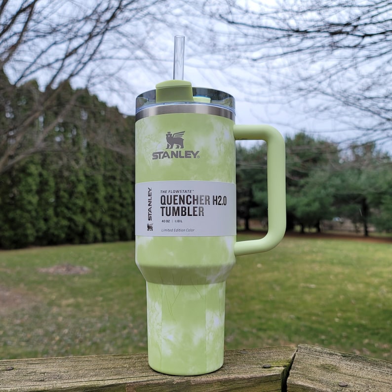 May include: A green and white tie-dye Stanley Quencher H2.0 tumbler with a handle and straw. The tumbler has a 40 oz capacity. The Stanley logo is visible on the front. The tumbler is a limited edition color.