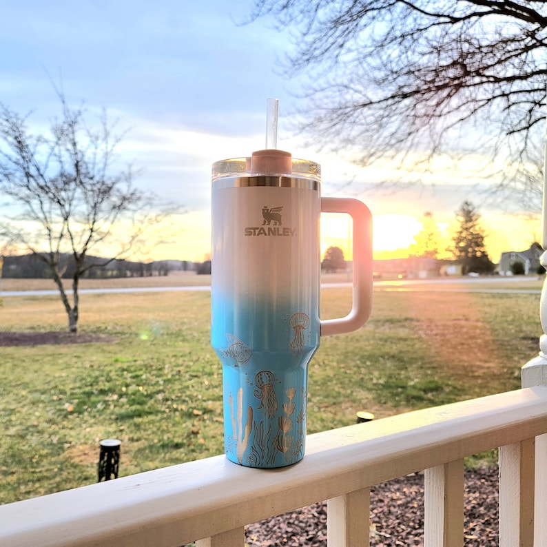 May include: A white and blue ombre Stanley tumbler with a handle and straw. The tumbler features a gold jellyfish and seaweed design. The Stanley logo is visible on the front. The tumbler is sitting on a white railing.