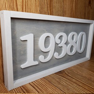 May include: A rectangular wooden sign featuring the number "19380" in white, raised lettering. The numbers are set against a grey background, framed by a white border. The sign is displayed on a wooden surface.