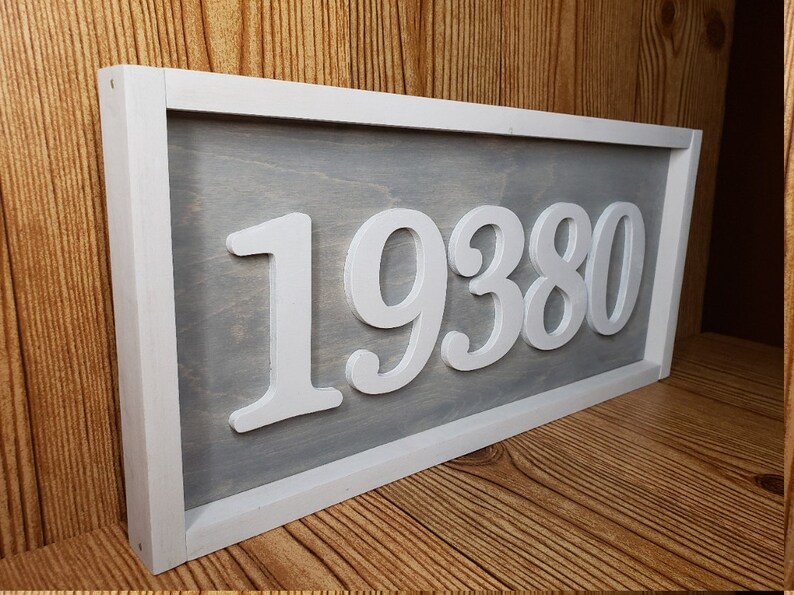 May include: A rectangular wooden sign featuring the number "19380" in white, raised lettering. The numbers are set against a grey background, framed by a white border. The sign is displayed on a wooden surface.
