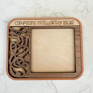 May include: Wooden picture frame with a musical theme. The frame has a rectangular opening and is decorated with musical notes and the text "COMPOSING NOTEWORTHY IDEAS". The frame is made of light and dark brown wood.
