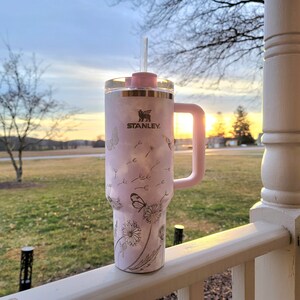 May include: A pink Stanley tumbler with a handle and straw, featuring a dandelion and butterfly design. The tumbler has the Stanley logo and a pink lid. The background shows a sunset over a grassy field.