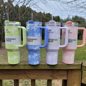 May include: Four Stanley Quencher H2.0 tumblers in pastel colors: green, blue, purple, and pink. Each tumbler has a matching handle and lid, with a clear straw. The tumblers have a tie-dye pattern and the text "QUENCHER H2.0 TUMBLER".