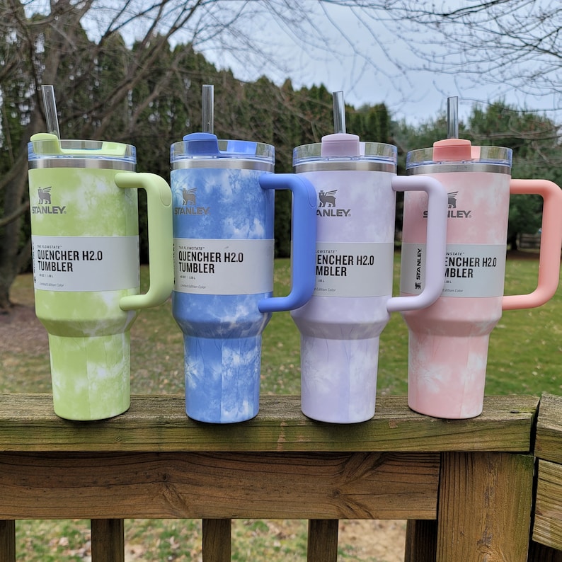 May include: Four Stanley Quencher H2.0 tumblers in pastel colors: green, blue, purple, and pink. Each tumbler has a matching handle and lid, with a clear straw. The tumblers have a tie-dye pattern and the text "QUENCHER H2.0 TUMBLER".
