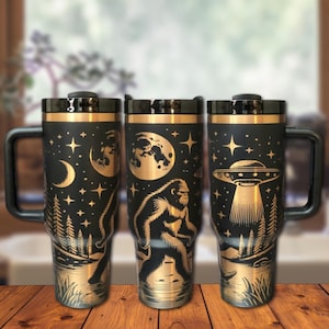 May include: Three stainless steel travel mugs with black and gold designs. The designs feature a crescent moon, a full moon, a UFO, a Bigfoot, and stars. The mugs have a black background and gold accents.