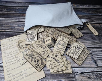 May include: A set of wooden tiles with illustrations of animals, including a bird, a fish, a rabbit, a cat, and a dog. The tiles are in a white fabric bag with the words "Tails & Tiles" printed on it. The tiles are part of a game called "Tails & Tiles" and the instructions for the game are printed on a piece of paper.