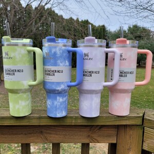 May include: Four Stanley Quencher H2.0 tumblers in pastel colors: green, blue, purple, and pink. Each tumbler has a matching handle and lid, with a clear straw. The tumblers are on a wooden deck.