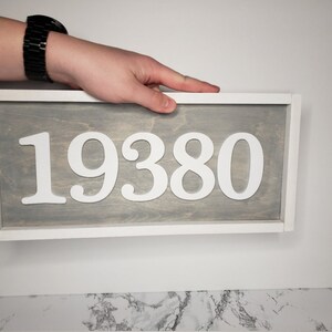 May include: A rectangular wooden sign displaying the number "19380" in white against a grey backdrop, framed in white. The sign is being held by a person.