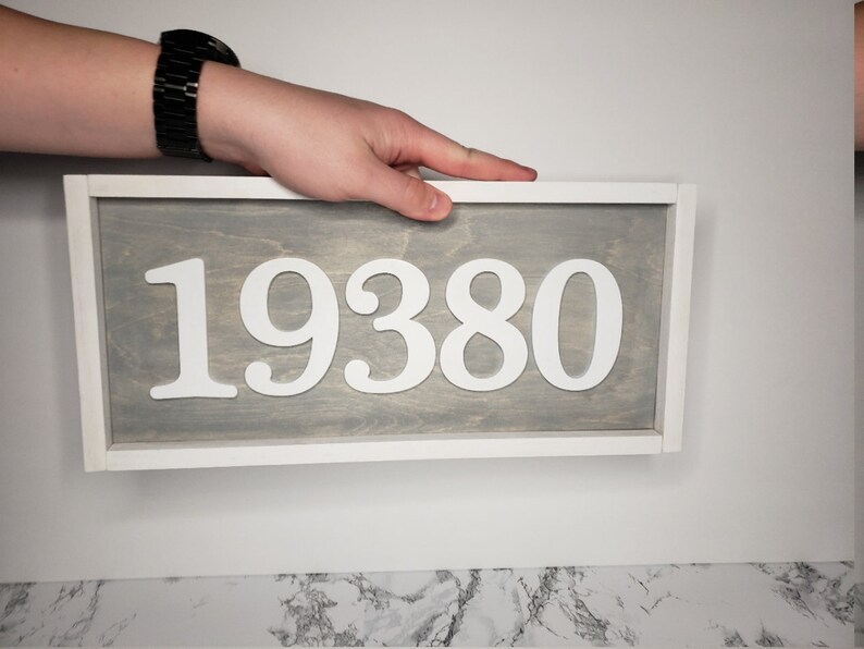 May include: A rectangular wooden sign displaying the number "19380" in white against a grey backdrop, framed in white. The sign is being held by a person.