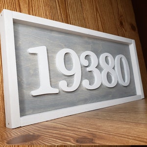 May include: A rectangular wooden sign with a white frame and a grey interior. The sign displays the number "19380" in white, three-dimensional lettering. The sign is resting on a wooden shelf.