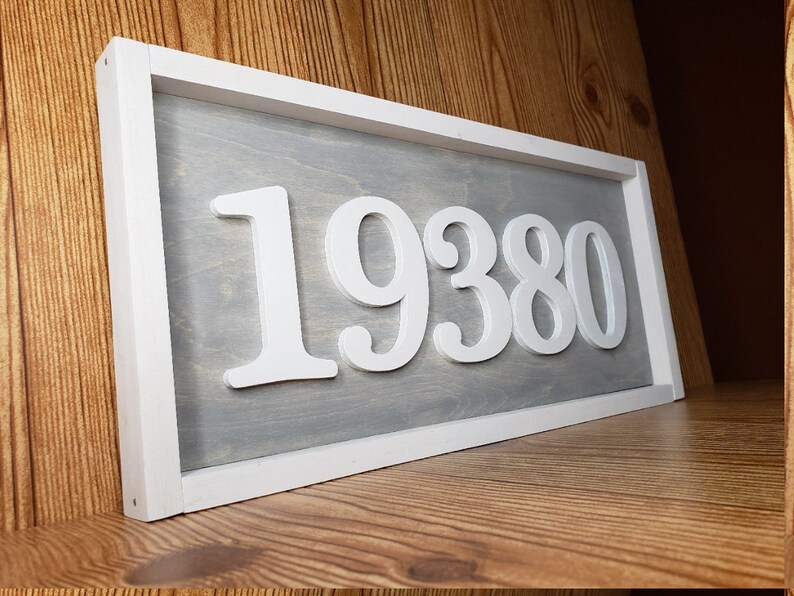 May include: A rectangular wooden sign with a white frame and a grey interior. The sign displays the number "19380" in white, three-dimensional lettering. The sign is resting on a wooden shelf.