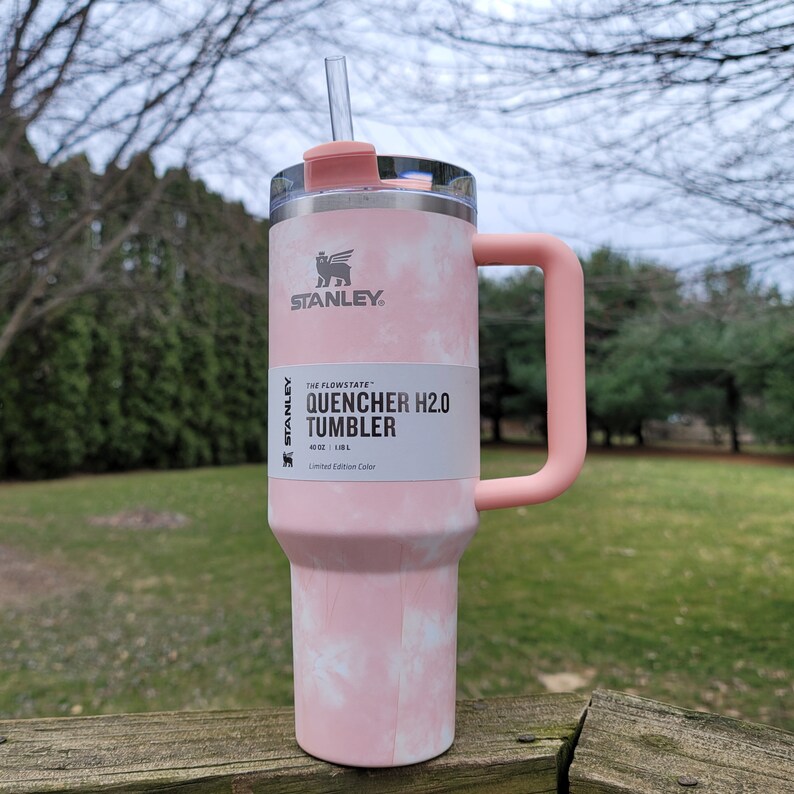 May include: A pink and white tie-dye Stanley Quencher H2.0 Tumbler with a handle and straw. The tumbler has a capacity of 40 oz (1.18 L) and features the Stanley logo. The tumbler is a limited edition color.