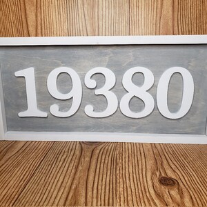 May include: A rectangular wooden sign displaying the number "19380" in white against a grey backdrop. The sign is framed in white and is placed on a wooden surface. This sign is suitable for home decor.