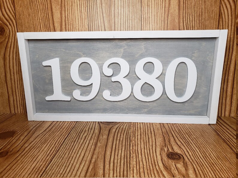May include: A rectangular wooden sign displaying the number "19380" in white against a grey backdrop. The sign is framed in white and is placed on a wooden surface. This sign is suitable for home decor.