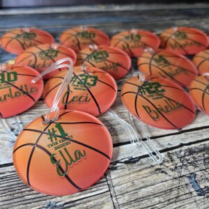 May include: A set of orange and black basketball-themed luggage tags with green text. Each tag has a number and a name, such as "#10 Charlotte", "#33 Ceci", and "#22 Briella". The tags are decorated with a basketball design and the text "Twin Valley Raiders".