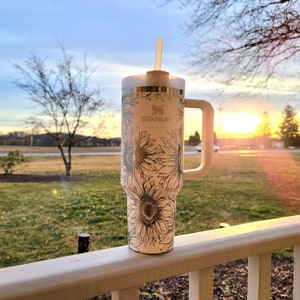 May include: A white Stanley tumbler with a sunflower design, a handle, and a straw. The tumbler is set against a backdrop of a sunset over a grassy field. The Stanley logo is visible on the tumbler.