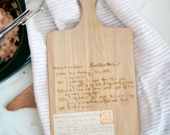 May include: A light-colored wooden cutting board with a handle, featuring handwritten recipe notes. The board rests on a white and gray striped cloth. The recipe is for raisin-filled cookies.