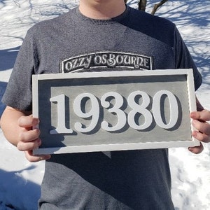 May include: A rectangular address sign displaying the number 19380 in white against a grey backdrop, framed in white. The sign is held by a person wearing a grey t-shirt. The text "OZZY OSBOURNE" is visible on the shirt.