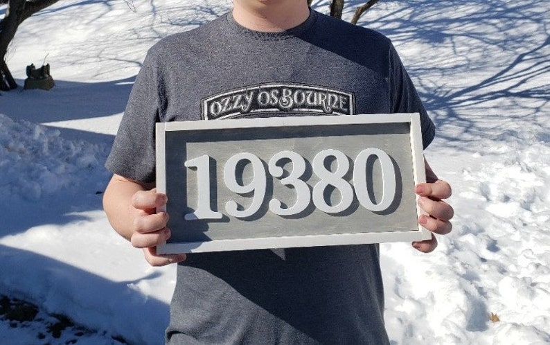 May include: A rectangular address sign displaying the number 19380 in white against a grey backdrop, framed in white. The sign is held by a person wearing a grey t-shirt. The text "OZZY OSBOURNE" is visible on the shirt.