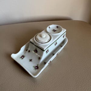 May include: White porcelain inkwell with two wells and a tray. The inkwell is decorated with a floral pattern of small pink roses and green leaves.