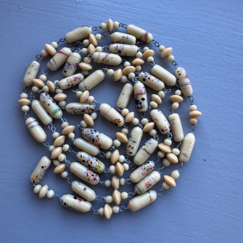 May include: A long necklace made of cream-coloured glass beads with colourful speckles. The beads are interspersed with smaller, round, cream-coloured beads. The necklace is strung on a silver chain.