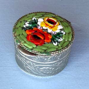 Op de afbeelding: Een rond zilverkleurig metalen doosje met een deksel versierd met een bloemrijk micro-mozaïekpatroon. Het mozaïek toont rode, gele en witte bloemen tegen een groene achtergrond. De zijkanten van het doosje zijn voorzien van een reliëfbladmotief.