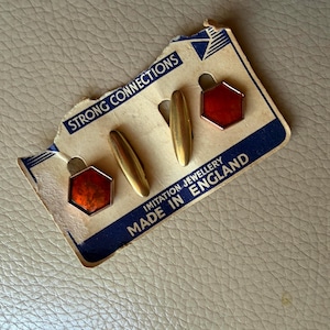 May include: Vintage gold-tone cufflinks and matching hexagon-shaped studs with red enamel centers. The cufflinks and studs are displayed on a torn cardboard backing with the text "STRONG CONNECTIONS" and "IMITATION JEWELLERY MADE IN ENGLAND".